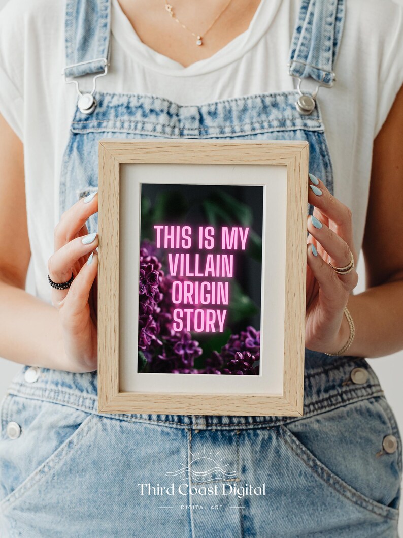 May include: A framed print with the text "THIS IS MY VILLAIN ORIGIN STORY" in pink neon against a dark background with purple flowers. The frame is light brown, and the image is held by a person wearing denim overalls.