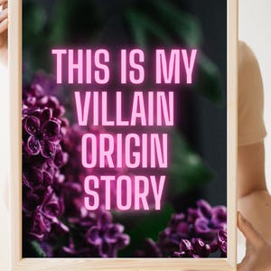 May include: A framed art print with the text "THIS IS MY VILLAIN ORIGIN STORY" in bright pink letters. The background features a blurred image of purple flowers. The frame is made of light-colored wood.