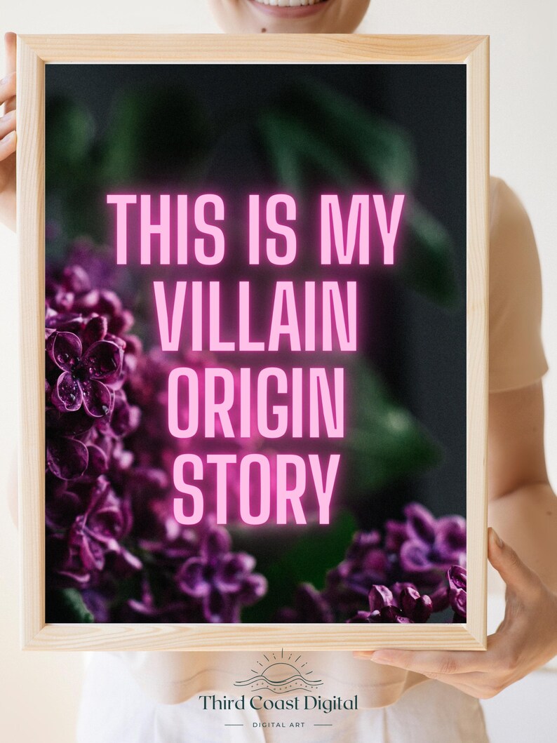 May include: A framed art print with the text "THIS IS MY VILLAIN ORIGIN STORY" in bright pink letters. The background features a blurred image of purple flowers. The frame is made of light-colored wood.
