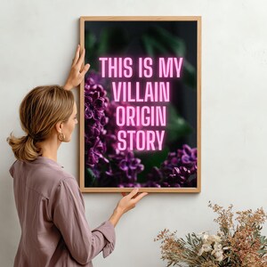 May include: A framed art print with the text "THIS IS MY VILLAIN ORIGIN STORY" in pink neon lettering. The artwork features a dark background with purple flowers. The frame is made of light-colored wood.
