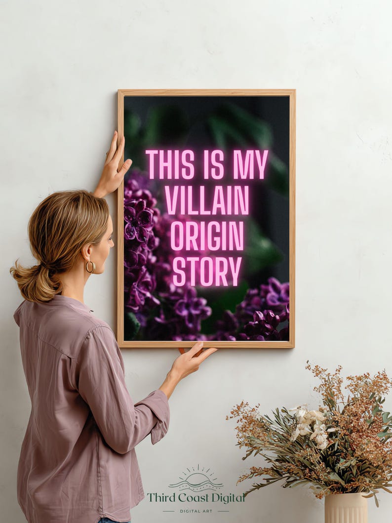 May include: A framed art print with the text "THIS IS MY VILLAIN ORIGIN STORY" in pink neon lettering. The artwork features a dark background with purple flowers. The frame is made of light-colored wood.