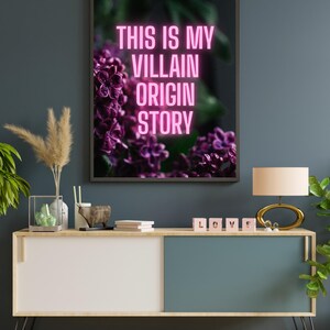 May include: A framed art print with the text "THIS IS MY VILLAIN ORIGIN STORY" in neon pink. The artwork features a close-up of purple flowers. The print hangs above a modern cabinet with a lamp and decorative items.