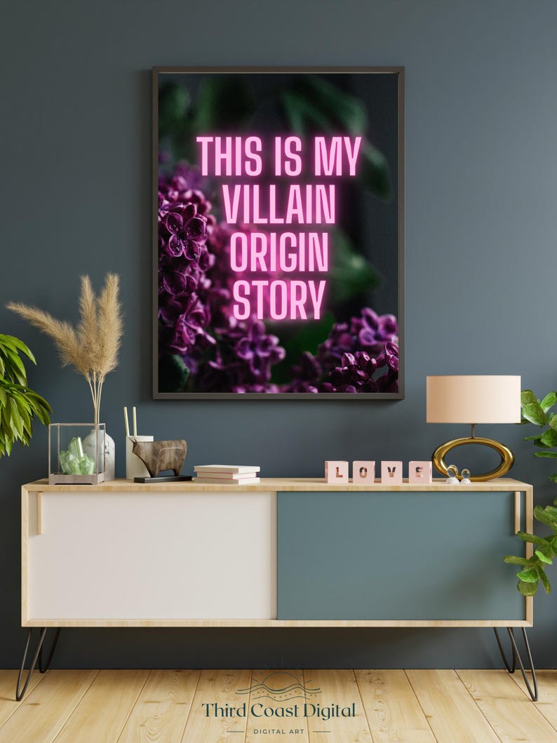 May include: A framed art print with the text "THIS IS MY VILLAIN ORIGIN STORY" in neon pink. The artwork features a close-up of purple flowers. The print hangs above a modern cabinet with a lamp and decorative items.