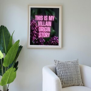 May include: Framed art print with the text "THIS IS MY VILLAIN ORIGIN STORY" in pink neon against a dark floral background. The print is in a white frame, displayed in a room with a white chair and a potted plant.