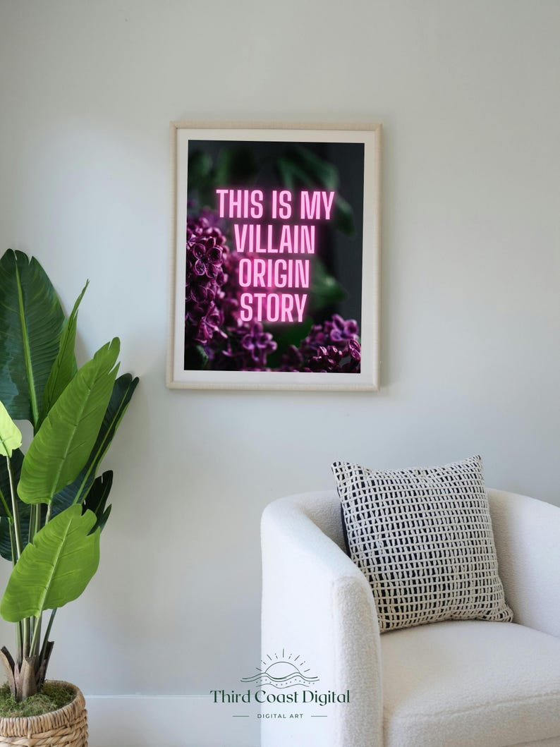 May include: Framed art print with the text "THIS IS MY VILLAIN ORIGIN STORY" in pink neon against a dark floral background. The print is in a white frame, displayed in a room with a white chair and a potted plant.