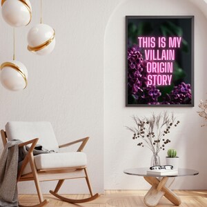 May include: A framed art print with the text "THIS IS MY VILLAIN ORIGIN STORY" in pink neon against a backdrop of purple flowers. The print hangs above a modern living room scene with a rocking chair, coffee table, and decorative lighting.