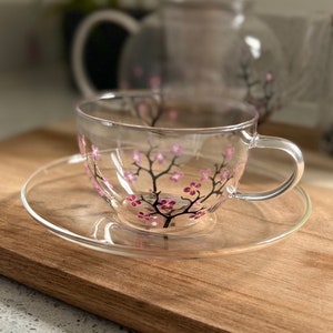 Könnte beinhalten: Ein klares Glas-Teetassen- und Untertassen-Set mit handgemaltem Design eines rosa Kirschblütenbaums. Das Set befindet sich auf einer Holzoberfläche.