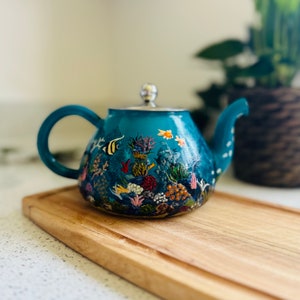 May include: A teal blue teapot with a hand-painted design of an underwater scene. The teapot features coral, fish, and other sea life.