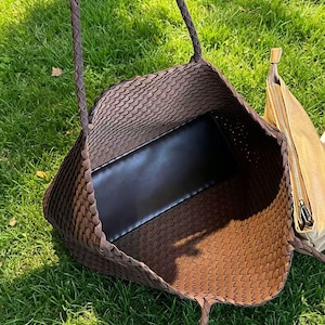 May include: A brown woven tote bag with braided handles, showcasing a spacious interior with a black base. A smaller, tan-colored zippered pouch is visible on the side. The bag is set against a backdrop of green grass.
