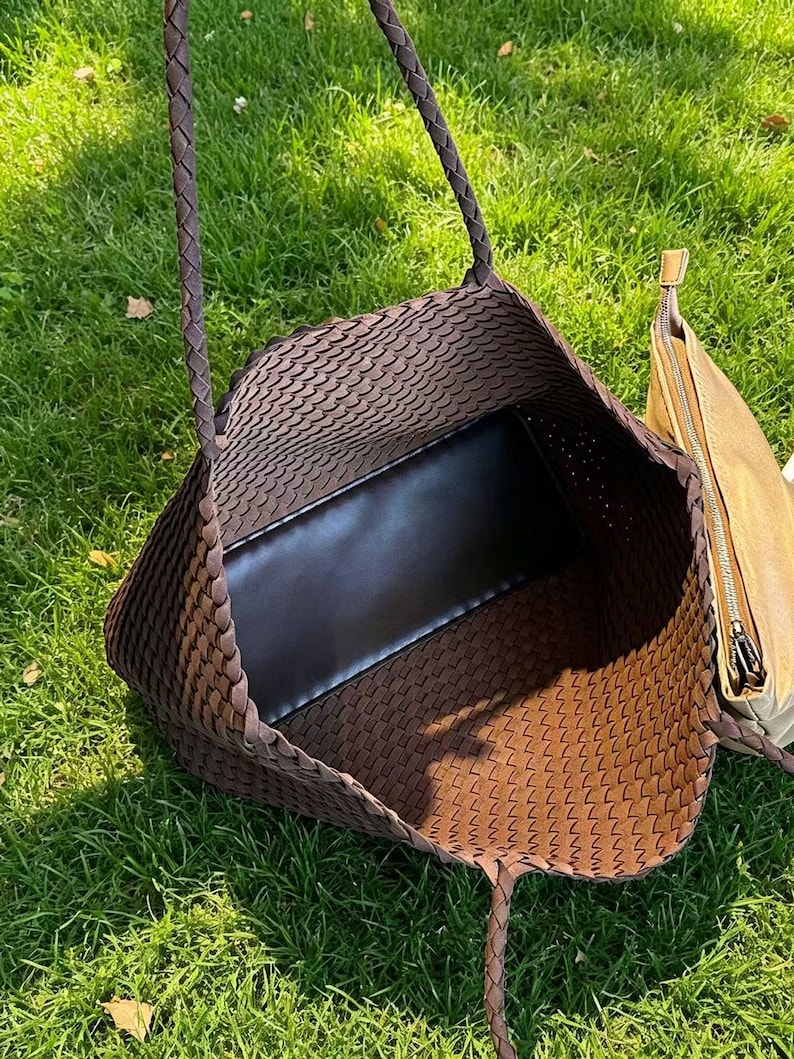May include: A brown woven tote bag with braided handles, showcasing a spacious interior with a black base. A smaller, tan-colored zippered pouch is visible on the side. The bag is set against a backdrop of green grass.