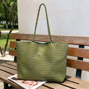 May include: A woven green tote bag with braided handles. The bag is made of a textured material and has a rectangular shape. The bag is sitting on a wooden bench, with a magazine visible at the bottom.