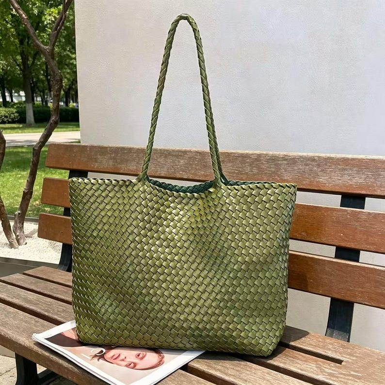 May include: A woven green tote bag with braided handles. The bag is made of a textured material and has a rectangular shape. The bag is sitting on a wooden bench, with a magazine visible at the bottom.