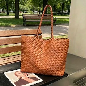 May include: A woven brown leather tote bag with braided handles. The bag is rectangular with a wide opening and sits on a black surface next to a magazine. The bag is made of a tightly woven pattern.