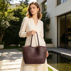 May include: A dark brown woven tote bag with braided handles. The bag has a rectangular shape and a spacious interior. The bag is held by a person wearing a cream-colored dress. The background features greenery and a building.