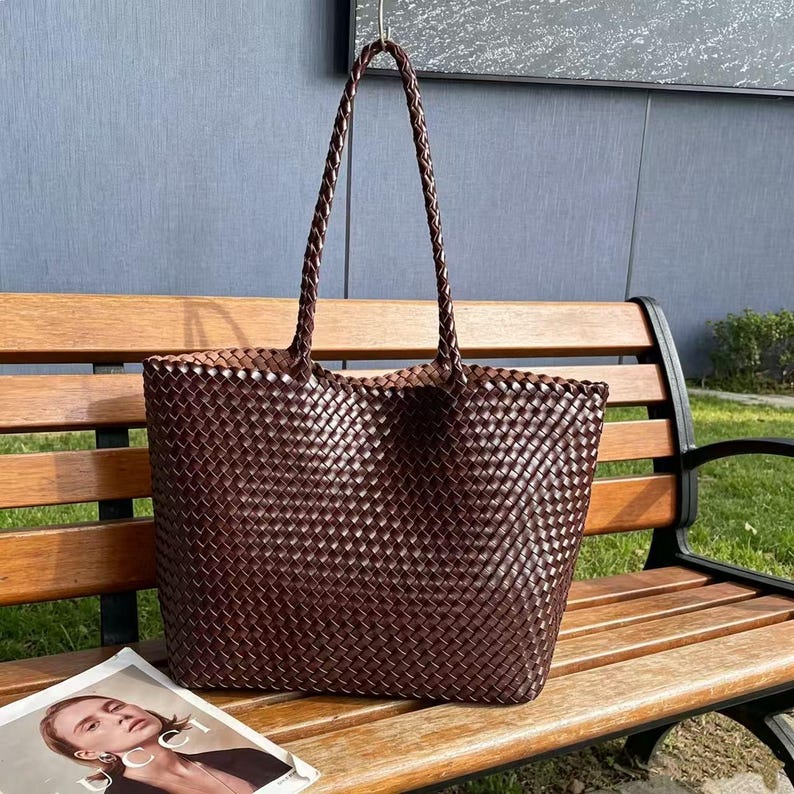 May include: A large, woven brown tote bag with braided handles. The bag is made of a tightly woven material, creating a textured surface. The bag is sitting on a wooden bench next to a magazine.