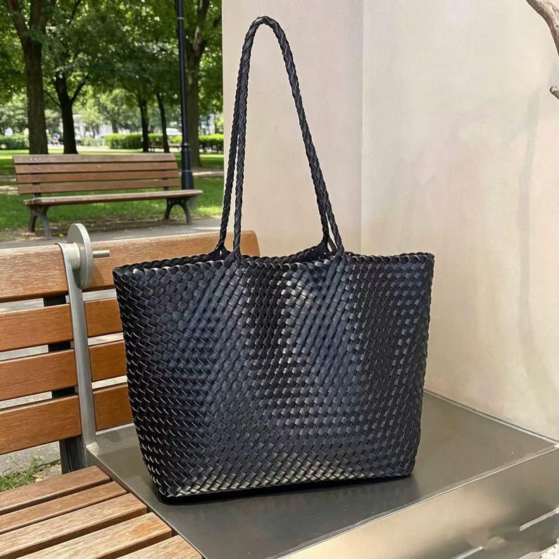 May include: A black woven tote bag with braided handles. The bag is rectangular with a wide opening and a textured, basket-weave design. The bag is sitting on a metal surface, with a park bench and trees in the background.