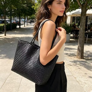 May include: A large, black woven tote bag with braided handles. The bag has a rectangular shape and a textured surface. The bag is carried over the shoulder. The bag is a stylish accessory.