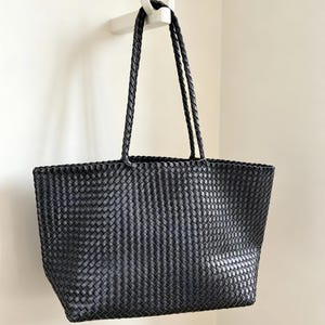 May include: A black tote bag with a woven design, hanging from a white hook on a white wall. The bag has two braided handles and a rectangular shape. The woven pattern gives the bag a textured appearance.