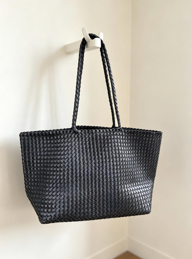 May include: A black tote bag with a woven design, hanging from a white hook on a white wall. The bag has two braided handles and a rectangular shape. The woven pattern gives the bag a textured appearance.