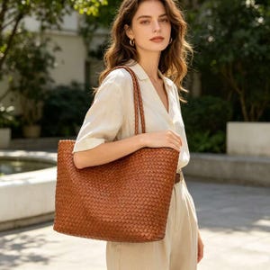 May include: A large, woven brown tote bag with braided handles. The bag has a rectangular shape and a textured surface. The bag is carried over the shoulder. The bag is made of a leather-like material.