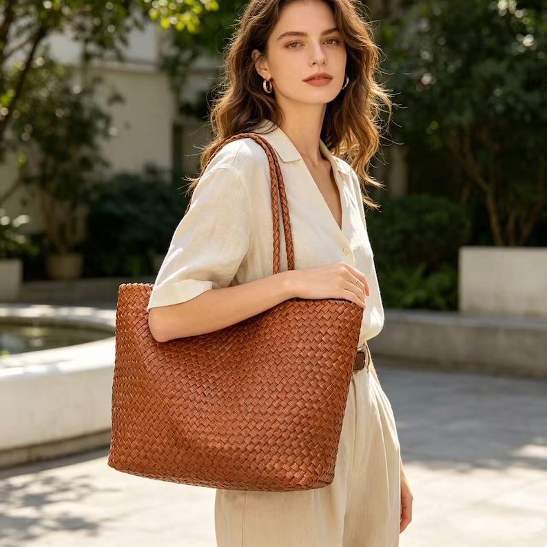 May include: A large, woven brown tote bag with braided handles. The bag has a rectangular shape and a textured surface. The bag is carried over the shoulder. The bag is made of a leather-like material.