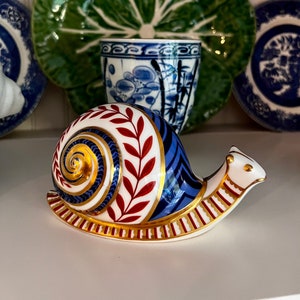 May include: A porcelain snail figurine with a blue, red, and gold painted shell. The shell has a spiral pattern and a leafy design. The snail is on a white surface.