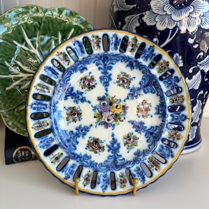 May include: A decorative, round, ceramic plate with a blue and white floral design. The plate has a pierced rim and a gold-colored edge. The plate is displayed on a gold-colored stand. A green plate and a blue and white vase are in the background.