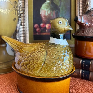May include: A ceramic pheasant-shaped dish with a brown and green glaze. The dish has a lid and is decorated with a detailed pheasant design. The dish is sitting on a brown surface.