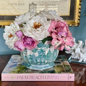 May include: A floral arrangement featuring white roses, pink hydrangeas, and purple accents in a blue and white pagoda-style vase. The vase sits atop a book titled "Palm Beach Living."