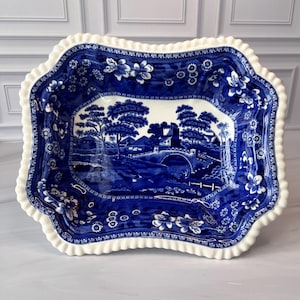 May include: A blue and white ceramic serving dish with a ruffled edge. The dish features a landscape scene in the center and floral patterns around the rim. The dish is rectangular with a scalloped edge.