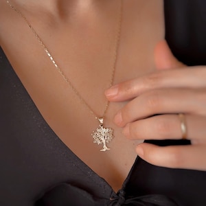 May include: A gold-colored necklace featuring a tree pendant. The tree pendant has detailed branches and a textured appearance. The necklace is worn against a dark background, highlighting the pendant's design. The chain is delicate and complements the pendant.