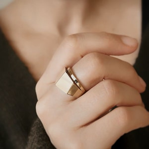 Könnte beinhalten: Ein goldfarbener Ring mit geometrischem Design. Der Ring hat eine rechteckige Oberseite und eine glatte, polierte Oberfläche. Der Ring wird am Finger getragen und unterstreicht seinen modernen und minimalistischen Stil. Das Design des Rings ist schlicht, aber elegant und für den Alltag geeignet.