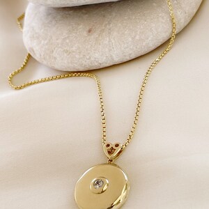 K&ouml;nnte beinhalten: Eine goldene Kettenkette mit einem runden Anh&auml;nger in Form eines Medallions, das einen kleinen, klaren Stein in der Mitte tr&auml;gt. Das Medaillon ist mit einem herzf&ouml;rmigen Verschluss an der Kette befestigt.