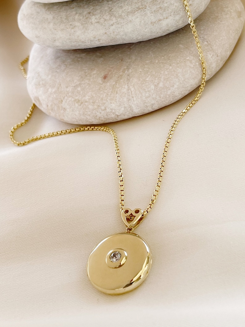K&ouml;nnte beinhalten: Eine goldene Kettenkette mit einem runden Anh&auml;nger in Form eines Medallions, das einen kleinen, klaren Stein in der Mitte tr&auml;gt. Das Medaillon ist mit einem herzf&ouml;rmigen Verschluss an der Kette befestigt.