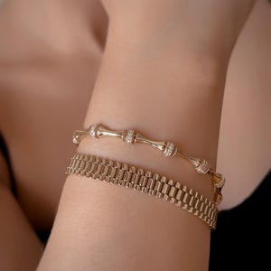 May include: Two gold bracelets on a person's wrist. The top bracelet is a delicate chain with small round stones. The bottom bracelet is a thicker chain with a more intricate design.