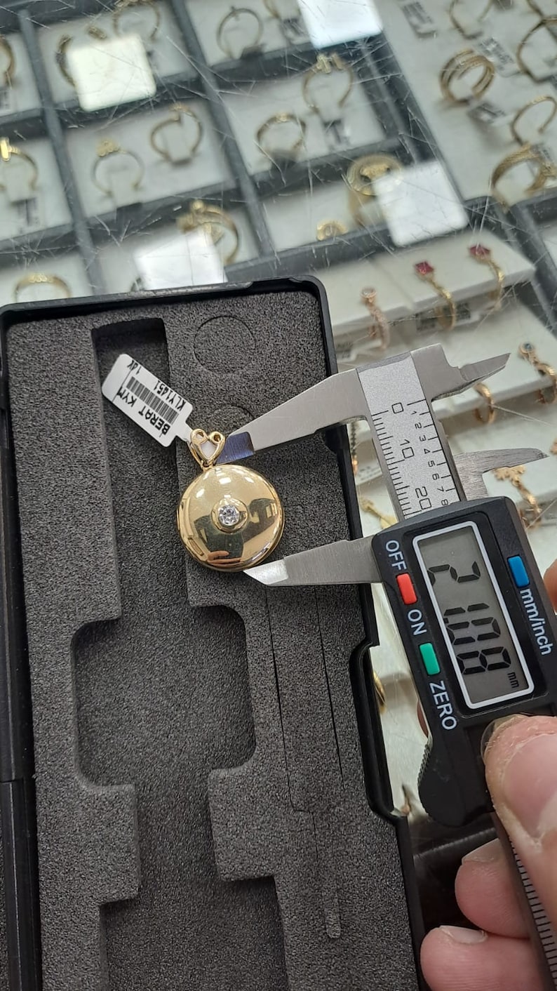 K&ouml;nnte beinhalten: Ein goldener Anh&auml;nger mit einem runden, flachen Design und einem einzigen klaren Stein in der Mitte. Der Anh&auml;nger wird mit einer digitalen Schieblehre gemessen, die 20,00 mm anzeigt.