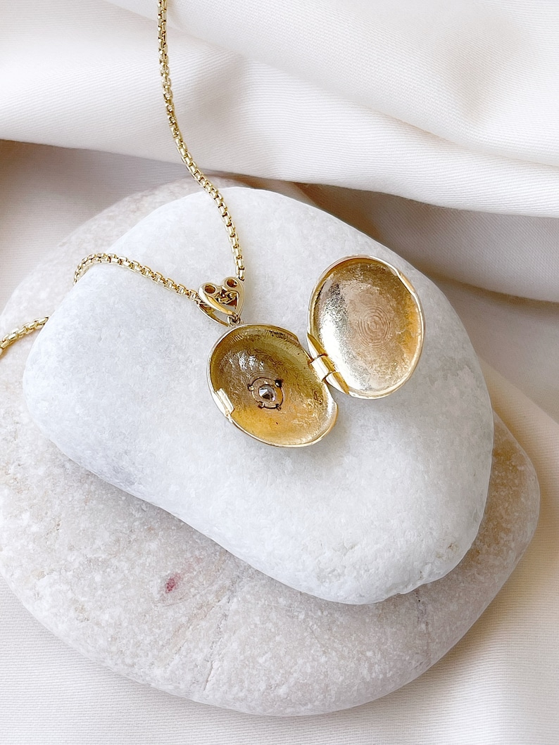 K&ouml;nnte beinhalten: Eine goldene Halskette mit einem Anh&auml;nger in Form eines Medaillons, mit einem herzf&ouml;rmigen Verschluss. Das Medaillon ist ge&ouml;ffnet und zeigt ein kleines, rundes, goldfarbenes Inneres.