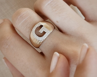 Può includere: Un anello d'oro con una grande lettera G incastonata con pietre di zircone cubico trasparenti.