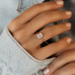 Pode incluir: Um anel de ouro com uma pedra central de diamante retangular com corte esmeralda. A pedra central é rodeada por um halo de diamantes menores. O anel é montado numa fina banda de ouro. O anel está no dedo de uma pessoa.