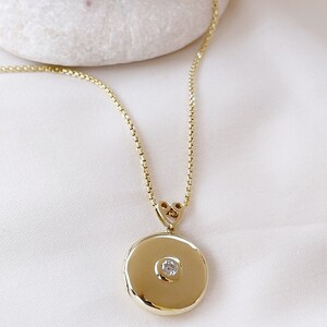K&ouml;nnte beinhalten: Eine goldene Kettenkette mit einem runden Anh&auml;nger in Form eines Medaillons. Das Medaillon hat einen kleinen, klaren Edelstein in der Mitte.