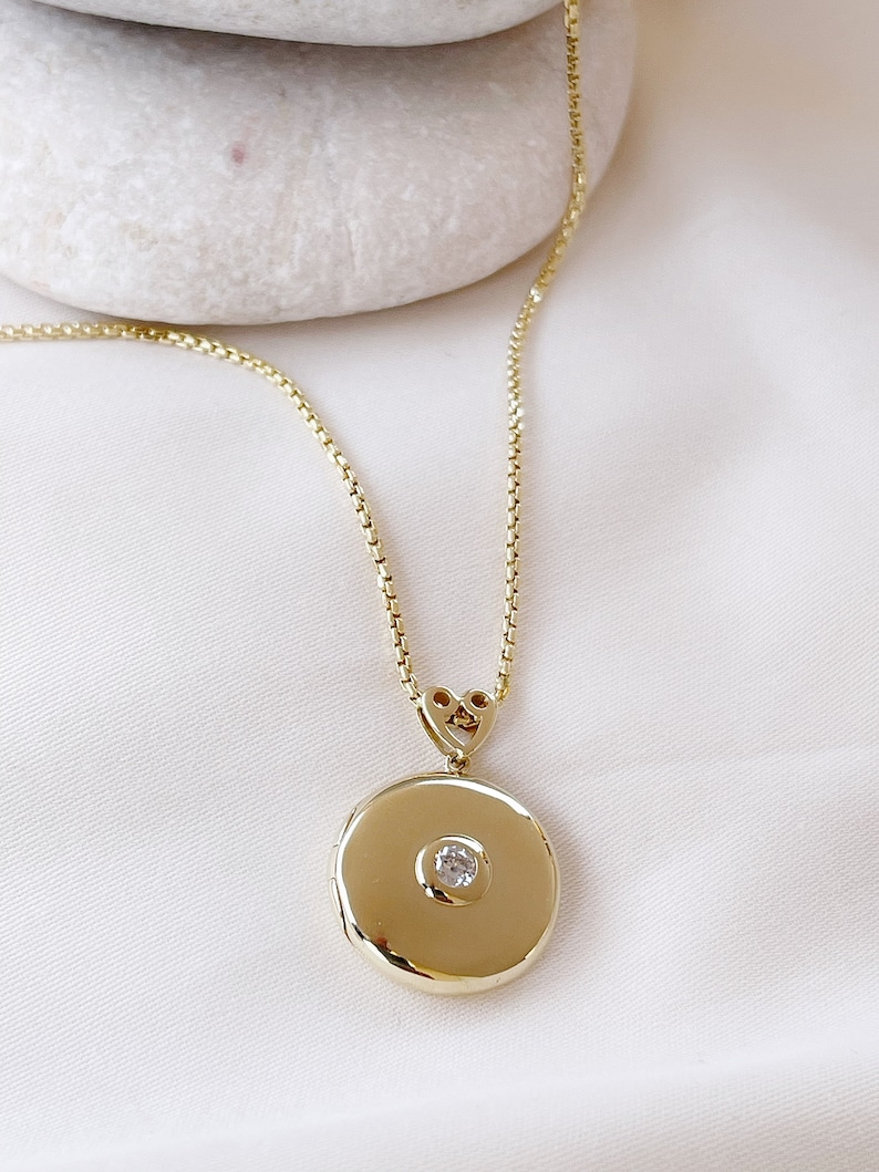 K&ouml;nnte beinhalten: Eine goldene Kettenkette mit einem runden Anh&auml;nger in Form eines Medaillons. Das Medaillon hat einen kleinen, klaren Edelstein in der Mitte.