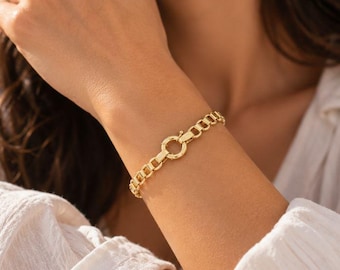 May include: A gold-toned chain bracelet featuring thick, rounded rectangular links and a prominent circular spring-ring clasp mechanism centered on the wrist. The metal has a polished, reflective finish that catches the light, creating a bright appearance.