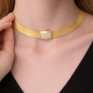May include: A gold-toned choker necklace with a rectangular, diamond-studded clasp. The necklace has a woven design and sits around the neck. The clasp is centered and adds a touch of sparkle to the piece. The necklace is worn against a black top.