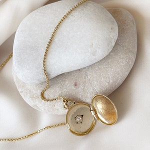 K&ouml;nnte beinhalten: Eine goldene Halskette mit einem kleinen Anh&auml;nger, der mit einem Diamanten besetzt ist. Der Anh&auml;nger ist ge&ouml;ffnet und zeigt ein leeres Inneres. Die Halskette liegt auf einem wei&szlig;en Hintergrund.