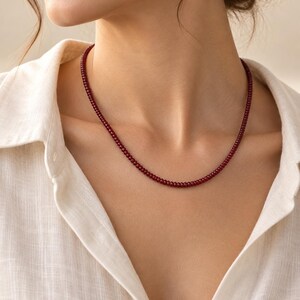 May include: A single strand necklace composed of small, uniform, deep red spherical beads. The necklace rests against the neck, featuring a consistent circular shape and a smooth, polished texture throughout the entire length of the beaded chain.