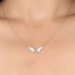 Pu&ograve; includere: Una collana con catena d'oro con un piccolo ciondolo delicato a forma di ali con un cerchio al centro. Le ali sono decorate con piccole pietre trasparenti.