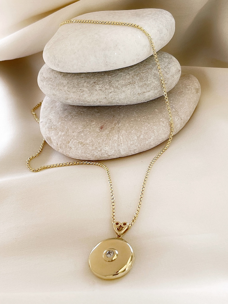 K&ouml;nnte beinhalten: Eine goldene Halskette mit einem runden Anh&auml;nger in Form eines Medaillons, verziert mit einem kleinen Diamanten. Die Kette liegt auf einem Stapel glatter, grauer Steine.