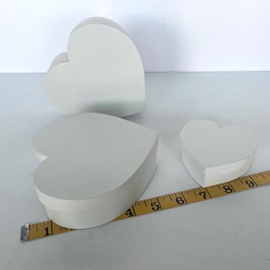 May include: Three white heart-shaped boxes of varying sizes. A yellow measuring tape is placed near the boxes. The largest box is approximately 4 inches wide. The boxes are made of a solid material and have a smooth, matte finish.
