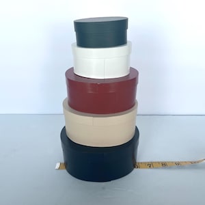 May include: A stack of five oval-shaped boxes in varying colors: dark green, white, burgundy, beige, and navy blue. A measuring tape is placed next to the boxes. The boxes are stacked on top of each other.