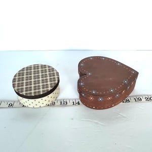 May include: Two decorative boxes. One is round with a brown and white plaid pattern on the lid and a cream-colored base with a small black cross pattern. The other is heart-shaped, brown, and decorated with small flowers around the edge.